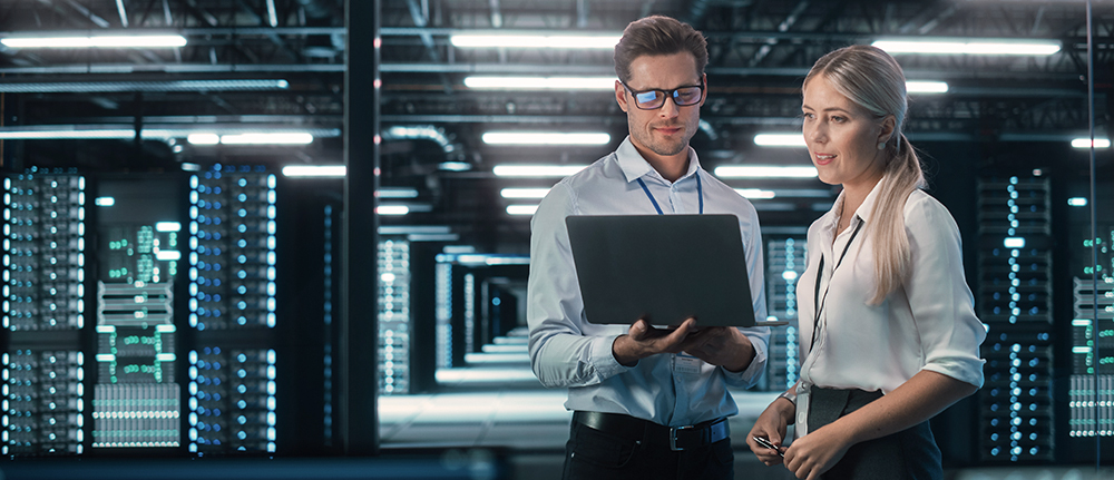 Two people standing in a server room next to large servers, while looking at a laptop together
