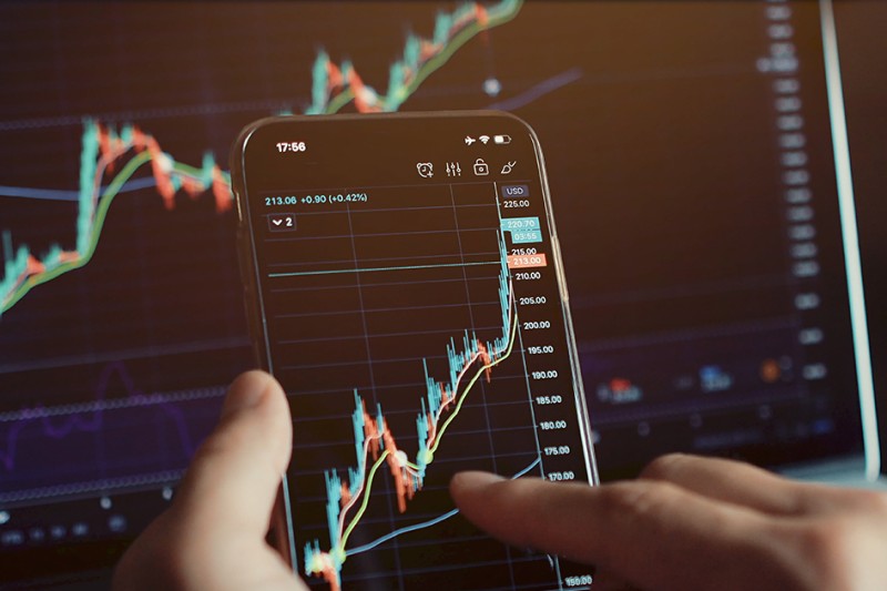 Close-up of a hand interacting with a smartphone displaying a financial chart with rising trends. A larger, similar chart is visible on a screen in the background.
